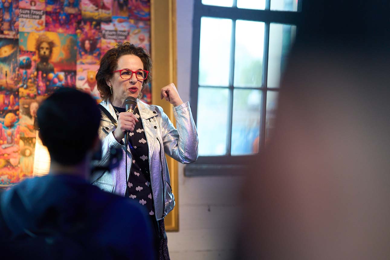 2024-fpc Alex, in a silver jacket, speaks in front of a collage and a window